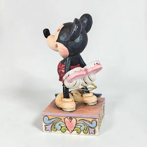 May include: A Mickey Mouse figurine, featuring a black head and ears, a red outfit, and tan shoes. The figurine is holding a pink object and stands on a decorative box with a heart design. The figurine is approximately 5 inches tall.