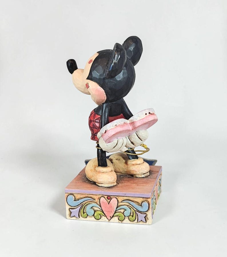May include: A Mickey Mouse figurine, featuring a black head and ears, a red outfit, and tan shoes. The figurine is holding a pink object and stands on a decorative box with a heart design. The figurine is approximately 5 inches tall.