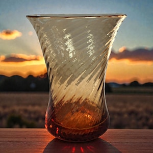 May include: A clear, amber-colored glass vase with a spiral design. The vase has a flared top and a rounded base. The background features a sunset with orange and blue hues, creating a warm and inviting atmosphere. The vase is sitting on a wooden surface.
