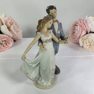 May include: A porcelain figurine of a dancing couple. The woman wears a white dress with pink accents and holds a bouquet. The man is in a grey suit with a red bow tie. The figurine is on a round base.