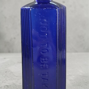 May include: A cobalt blue antique poison bottle with vertical ribbed sides. The rectangular bottle has a narrow neck and the words "NOT TO BE TAKEN" are embossed on the front. The bottle is made of glass.