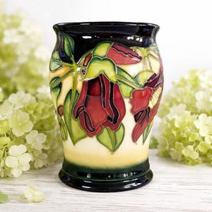May include: A ceramic vase with a glossy black rim and base, featuring a hand-painted floral design. The flowers are a deep red with green leaves and yellow accents. The vase is approximately 8 inches tall.