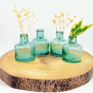 May include: Four small, teal blue glass bud vases with a textured surface, arranged on a wooden slice. The vases are filled with dried flowers and greenery.