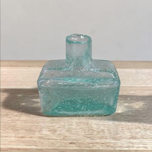 May include: A small, antique, square-shaped glass bottle with a light teal color. The bottle has a short, cylindrical neck and a textured surface. The bottle is sitting on a wooden surface.