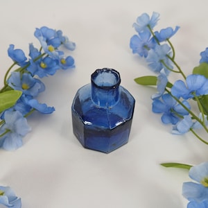 Peut inclure: Un petit encrier en verre bleu cobalt de forme octogonale. Le col de l'encrier est cylindrique. Il est posé sur une surface blanche, entouré de fleurs bleues à cœur jaune et de feuilles vertes. Objet vintage.