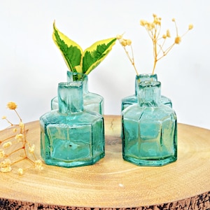 May include: Four teal blue glass bottles with a hexagonal shape and a rounded top. The bottles are arranged in a circle on a wooden surface. The bottles are empty and have a vintage look.
