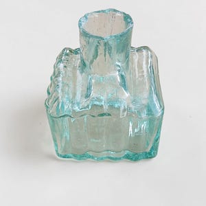 May include: A clear, turquoise glass bottle with a distinctive, textured design. The bottle features a rectangular base and a cylindrical neck. The glass has a slightly bubbly appearance, enhancing its vintage appeal. The bottle is likely a decorative item.