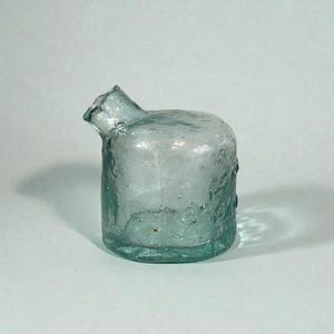 Peut inclure: Une bouteille en verre transparent de style ancien, avec un corps court et cylindrique et un bec incliné vers le haut. Le verre a une surface légèrement texturée et une subtile teinte bleu-vert, suggérant l'âge et l'utilisation. Il s'agit probablement d'un récipient d'apothicaire ou de laboratoire vintage.