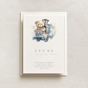 May include: A watercolour illustration of a teddy bear driving a blue train with white smoke. The text "LUCAS is turning three" is below the image. The invitation includes the date, time, location, and RSVP information.