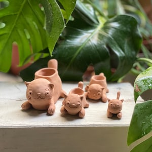 Könnte beinhalten: Vier terracotta Katzen-Pflanztopf in verschiedenen Größen. Der größte Katzen-Pflanztopf liegt mit ausgestreckten Pfoten. Die anderen drei Katzen-Pflanztöpfe sitzen aufrecht. Alle Katzen-Pflanztöpfe haben ein einfaches, handgemaltes Design.