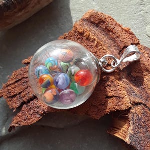 May include: A clear glass pendant filled with colorful marbles. The marbles are various colors including red, blue, green, orange, and purple. The pendant has a silver-colored bail for attaching to a chain.