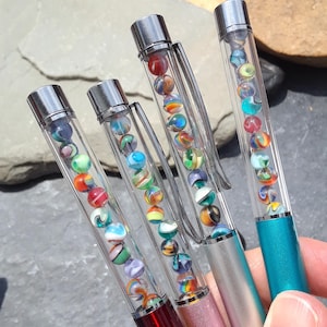 May include: Four pens with clear barrels filled with colorful marbles. The pens have silver accents and clips, with the pen bodies in red, pink, silver, and teal. The marbles are a mix of colors and patterns.