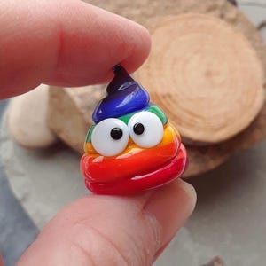 May include: A colorful, handcrafted glass figurine shaped like a smiling poop emoji. The figurine features a rainbow of colors, including red, orange, yellow, green, blue, and purple, with large white eyes and black pupils.