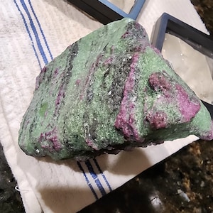 May include: A large, rough, green and pink gemstone with a rough, uneven surface. The gemstone has a mix of green and pink colors, with some black streaks.