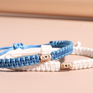 May include: Two matching friendship bracelets. One is blue, the other is white. Each bracelet has a gold-colored rectangular bead with initials and a heart symbol. The bracelets are made of woven cord.