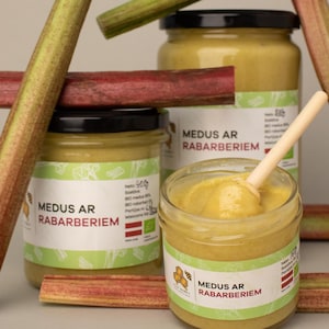 May include: A close-up shot of jars of honey with rhubarb. The jars have black lids and labels with text in Latvian. Rhubarb stalks in shades of green and red are arranged around the jars. A wooden spoon rests in one jar.