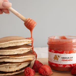 May include: A stack of pancakes with strawberry honey being drizzled from a wooden honey dipper. A jar of strawberry honey and fresh strawberries are next to the pancakes on a wooden board. The jar label reads "MEDUS AR ZEMENĒM".