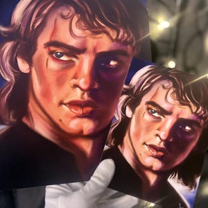 May include: Two art prints featuring a portrait of a man with light skin, blue eyes, and dark hair. The artwork has a painterly style with a focus on the face, showcasing a stern expression. The prints are held by a gloved hand.