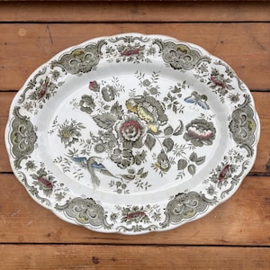 May include: Oval serving platter with a white base and a floral design in shades of green, grey, red, and yellow. The rim features a decorative border. A bird is depicted on the platter.