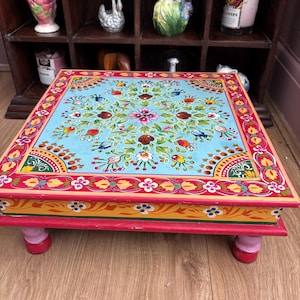 May include: A square wooden low table featuring a light blue top with a hand-painted floral pattern in red, green, and white. The border is decorated with red, yellow, and white motifs. The table has four short, rounded, pink and red legs.