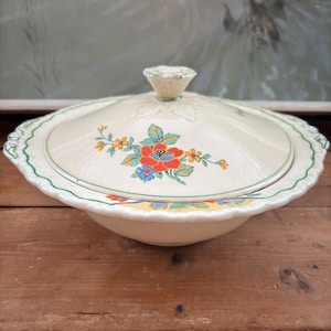 May include: A white ceramic serving bowl with a lid, featuring a floral design in shades of red, yellow, green, and blue. The bowl has a scalloped edge and a raised floral pattern on the lid.