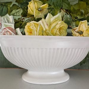 May include: A white, fluted ceramic bowl with a pedestal base. The bowl is filled with yellow roses, set against a backdrop of a floral painting. The bowl's design features vertical grooves.