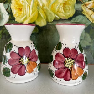 May include: A pair of white ceramic vases with a hand-painted floral design. Each vase has a burgundy flower with an orange accent and a yellow centre. Green leaves and vines surround the flowers. The rims are trimmed in burgundy.
