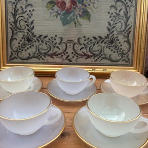 May include: A collection of six vintage teacups and saucers. The cups and saucers are white with gold edging. A framed floral needlepoint artwork is in the background.