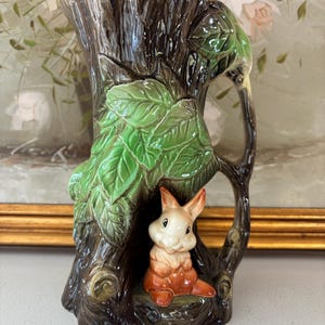 May include: A ceramic vase in the form of a tree trunk, featuring green leaves and a small orange and white bunny figurine. The vase has a handle and is painted in brown and green tones, creating a whimsical, nature-inspired design.