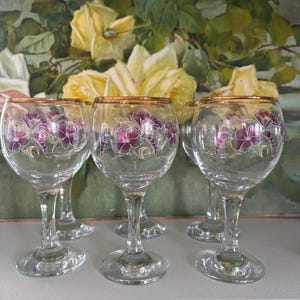 May include: A collection of six clear wine glasses, each with a gold rim and a purple floral pattern. The glasses are stemmed and arranged together. The backdrop is a painting of yellow roses and green leaves, creating a vintage aesthetic.