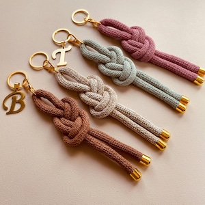 May include: Four keyrings with a knotted rope design in shades of brown, grey, green, and purple. Each keyring has a gold-coloured ring and a gold-coloured initial charm or end cap.