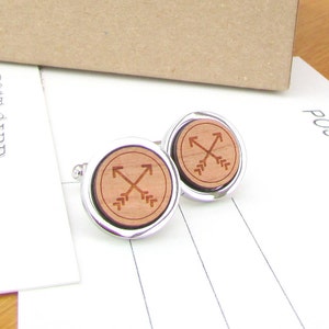 May include: A pair of silver cufflinks with a wooden inlay featuring a design of two crossed arrows. The arrows are brown and the wood is a light brown color.
