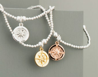 May include: Three silver beaded bracelets with compass charms. The charms are silver, gold, and rose gold.
