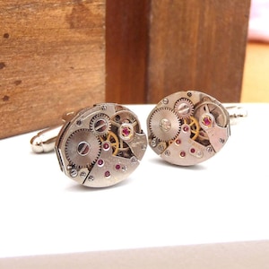 May include: A pair of silver cufflinks with a circular design featuring intricate clockwork gears and ruby-colored accents.