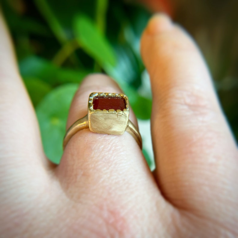 May include: A gold ring with a rectangular, reddish-brown gemstone. The ring has a brushed metal finish and a simple, elegant design. The gemstone is set in a bezel setting with a scalloped edge.
