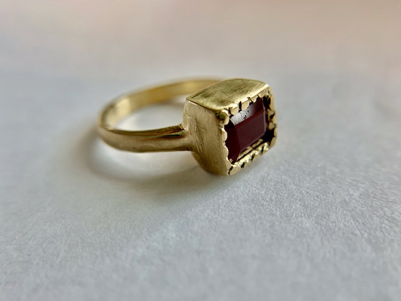 May include: A gold-colored ring with a rectangular, dark red gemstone. The ring has a textured band and a decorative, scalloped setting around the stone. The ring is shown in a close-up shot.