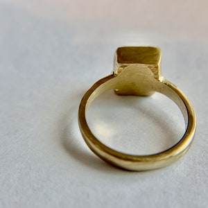 May include: A gold-colored ring with a square-shaped stone. The ring band is smooth and rounded, leading up to the square stone. The ring is set against a white background.