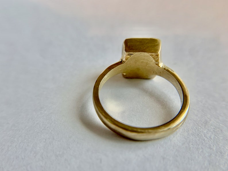 May include: A gold-colored ring with a square-shaped stone. The ring band is smooth and rounded, leading up to the square stone. The ring is set against a white background.