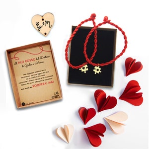 Può includere: Due braccialetti di corda rossa con ciondoli a forma di puzzle dorati in una scatola con un cuore rosso e il testo "A FILO ROSSO del Destino di Giulia e Marco" e il testo "ROMPERA' MAI".