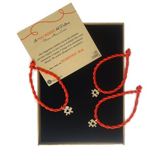 Può includere: Due braccialetti di filo rosso con ciondoli a forma di puzzle dorati. I braccialetti sono in una scatola nera con un biglietto che recita "Il filo rosso del destino" in italiano. Il biglietto spiega che un filo rosso invisibile collega le persone destinate a incontrarsi.