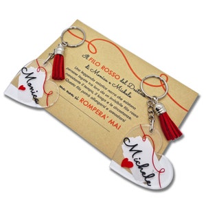 Può includere: Due portachiavi a forma di cuore con nappine rosse, ognuno con un cuore di carta bianca all'interno con i nomi "Monica" e "Michele" scritti in nero. I portachiavi sono accompagnati da una carta con il testo "A filo rosso del destino di Monica e Michele", che si traduce in "Un filo rosso del destino di Monica e Michele".