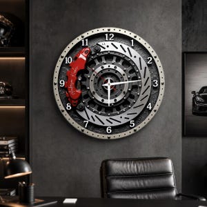 Könnte beinhalten: Eine runde Wanduhr, die wie eine Bremsscheibe mit Bremssattel gestaltet ist. Die Uhr verfügt über einen silberfarbenen Außenring mit schwarzen Ziffern, ein dunkel texturiertes Zentrum und einen roten Bremssattel mit weißer Schrift.
