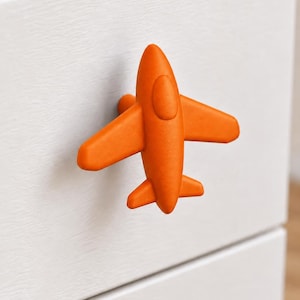 Op de afbeelding: Een feloranje, vliegtuigvormige ladeknop gemonteerd op een witte kastfront. De knop heeft een gladde, matte afwerking met gevormde details voor de cockpit, vleugels en staartsectie, en fungeert als een functionele handgreep voor laden.