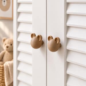 Op de afbeelding: Twee lichtbruine, bolvormige kastknoppen in de vorm van berenkoppen met kleine, ronde oortjes. Deze knoppen zijn bevestigd aan de voorzijde van een witte kastdeur met horizontale lamellen en bieden een grip voor het openen van de deuren.