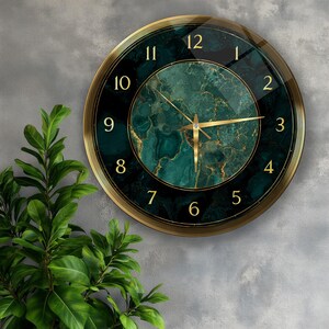 May include: A round wall clock with a gold-colored frame and hands. The clock face features a dark green marble design with gold accents and gold numerals. The clock is mounted on a gray wall, with green leafy plants in the foreground.