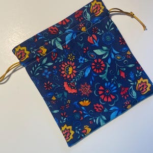 May include: A navy blue drawstring pouch with a vibrant floral pattern in red, yellow, and blue. The fabric features a repeating design of stylized flowers and leaves. The drawstring is a gold color.