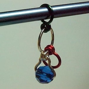 Pu&ograve; includere: Un ciondolo con una perla di cristallo blu con accenti di filo dorato e rosso pende da un anello di filo verde. Il ciondolo &egrave; attaccato a una barra di metallo argentato.