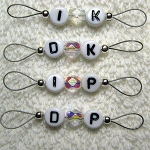 May include: Four sets of handmade stitch markers. Each marker has white letter beads spelling out different letter combinations, with a clear, iridescent bead in the center. The markers are attached to a silver wire loop.