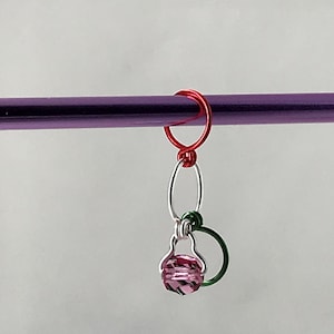 May include: A pink crystal bead hangs from a silver and green wire loop, attached to a red wire ring. The ring is attached to a purple knitting needle.