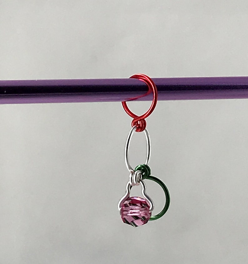 May include: A pink crystal bead hangs from a silver and green wire loop, attached to a red wire ring. The ring is attached to a purple knitting needle.